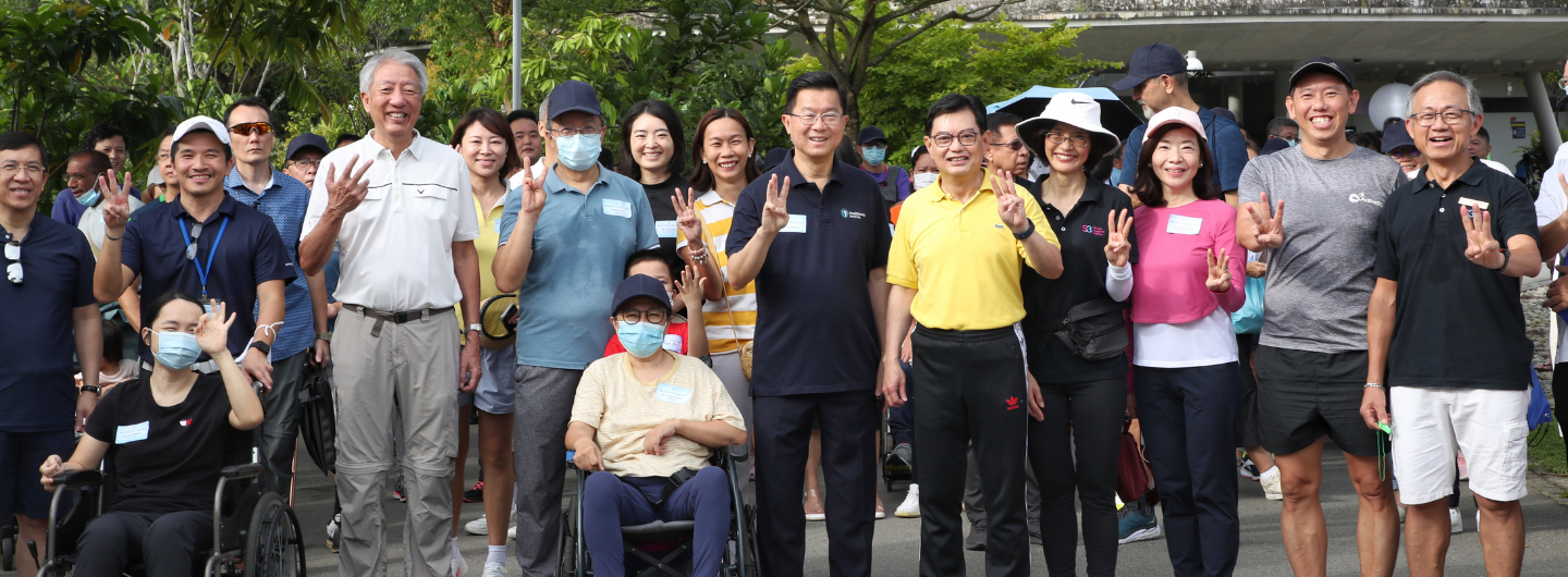 Group of people in casual wear outdoors, some in wheelchairs, raising three fingers.